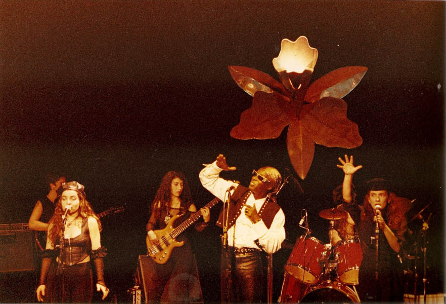 Fotografia colorida. Itamar Assumpção e Às integrantes da banda Orquídeas (Miria, Maria, Clara Bastos e Tata Fernandes) se apresentam em show. A imagem de uma orquídea de grandes dimensões faz parte do cenário.